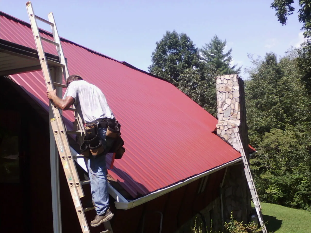 Roofer on a ladder beside a red metal roof and stone chimney — Soffit & Fascia Repair by Roofing Brothers in Middlebury Center